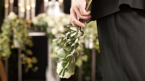 Mann in schwarzem Anzug hält einen Strauß mit grünen Blättern und einer weißen Rose vor einem dekorierten Hintergrund.